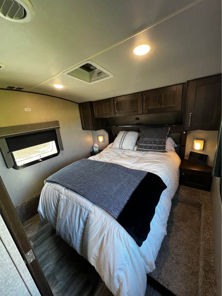 The front bedroom of The Reflection with a made-up queen bed between two nightstands, overhead cabinets above the headboard, a window alongside and a roof vent