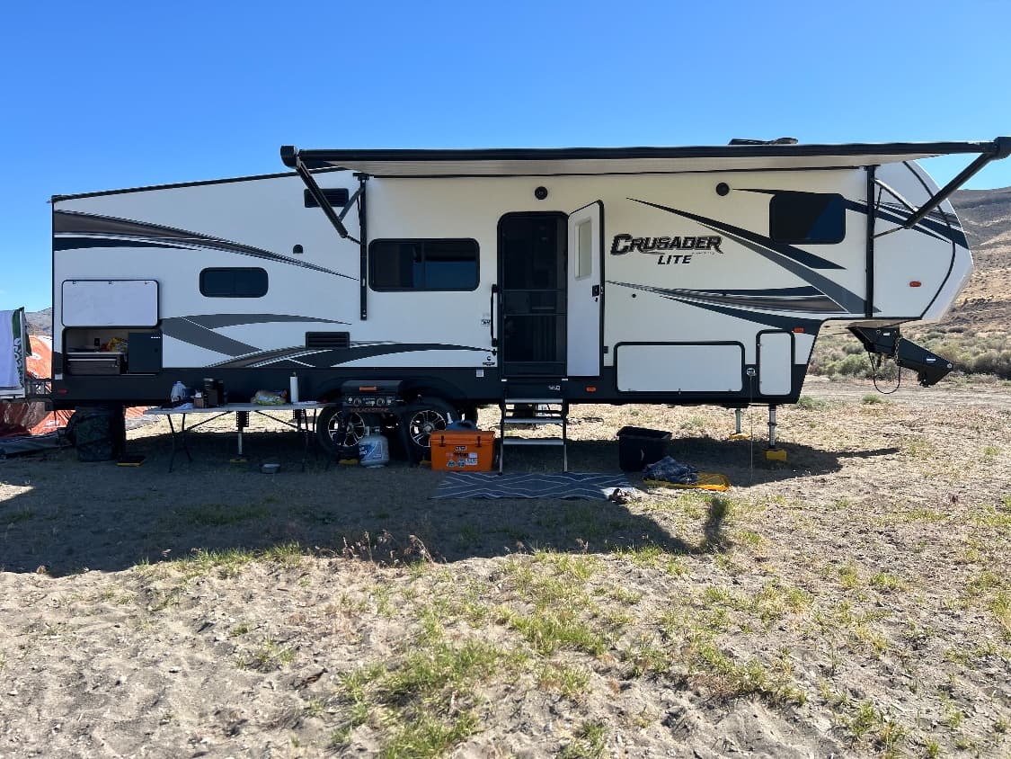 The Crusader, a cream and grey Crusader Lite fifth wheel with its patio awning extended and its outside kitchen open, set up on high desert scrub with hills behind it